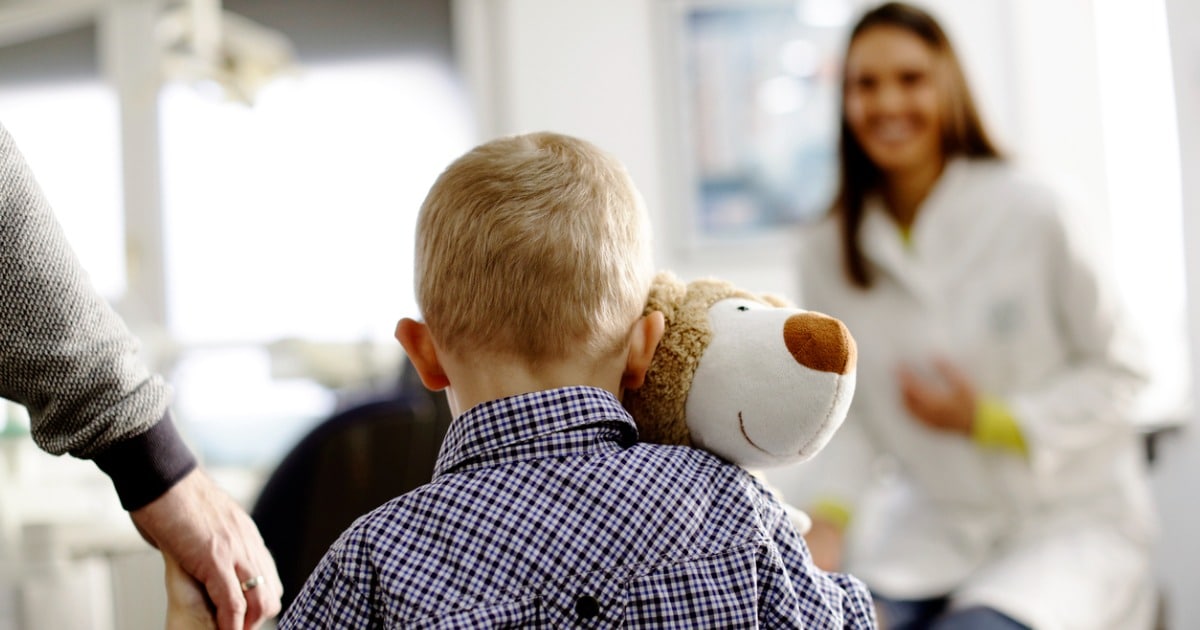 A smiling dental professional greets a parent or guardian who appear to be entering a clinic. We see the parent and child's backside. The boy is holding a stuffed animal.