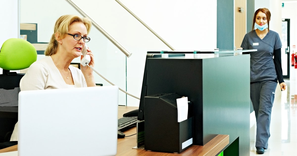 Worker is on the phone at the front desk of a dental or medical clinic. A dental assistant or hygienist wearing scrubs approaches in the background.