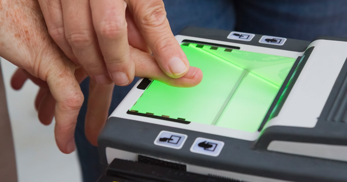 A person's hand appears to be guiding another person's finger on a digital fingerprint scanner such as a live scan fingerprint scanner.