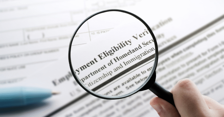 A magnifying glass is held over what appears to be a Form I-9. This text is readable: Employment Eligibility Verification, Department of Homeland Security, Citizenship and Immigration