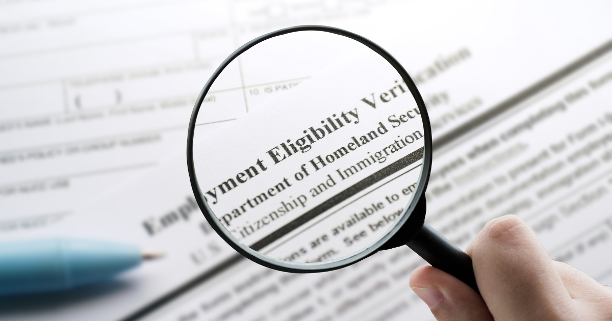 A magnifying glass is held over what appears to be a Form I-9. This text is readable: Employment Eligibility Verification, Department of Homeland Security, Citizenship and Immigration
