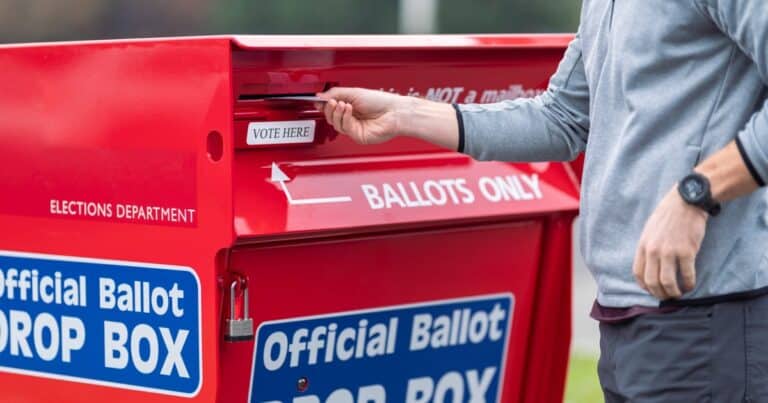 Man's hand is dropping a ballot into the slot of a ballot box. Visibly printed on the drop box are BALLOTS ONLY, Official Ballot Drop Box and Vote Here