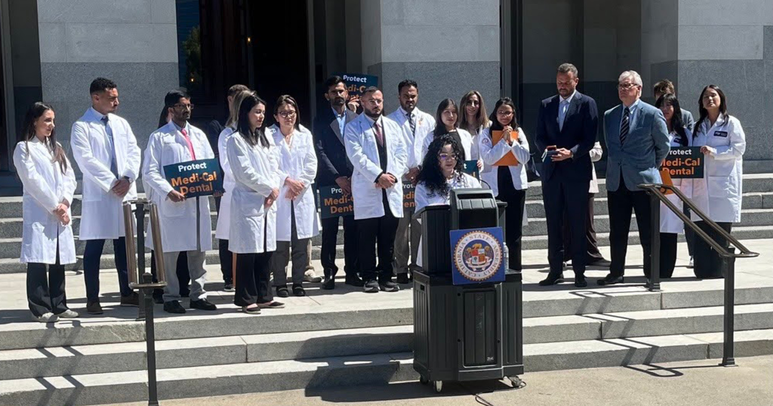 A group of dental students in their white coats stand behind a speaker at a podium on the Capitol steps. Some students are holding signs that read "Protect Medi-Cal Dental." An emblem on the podium reads "California State Assembly"