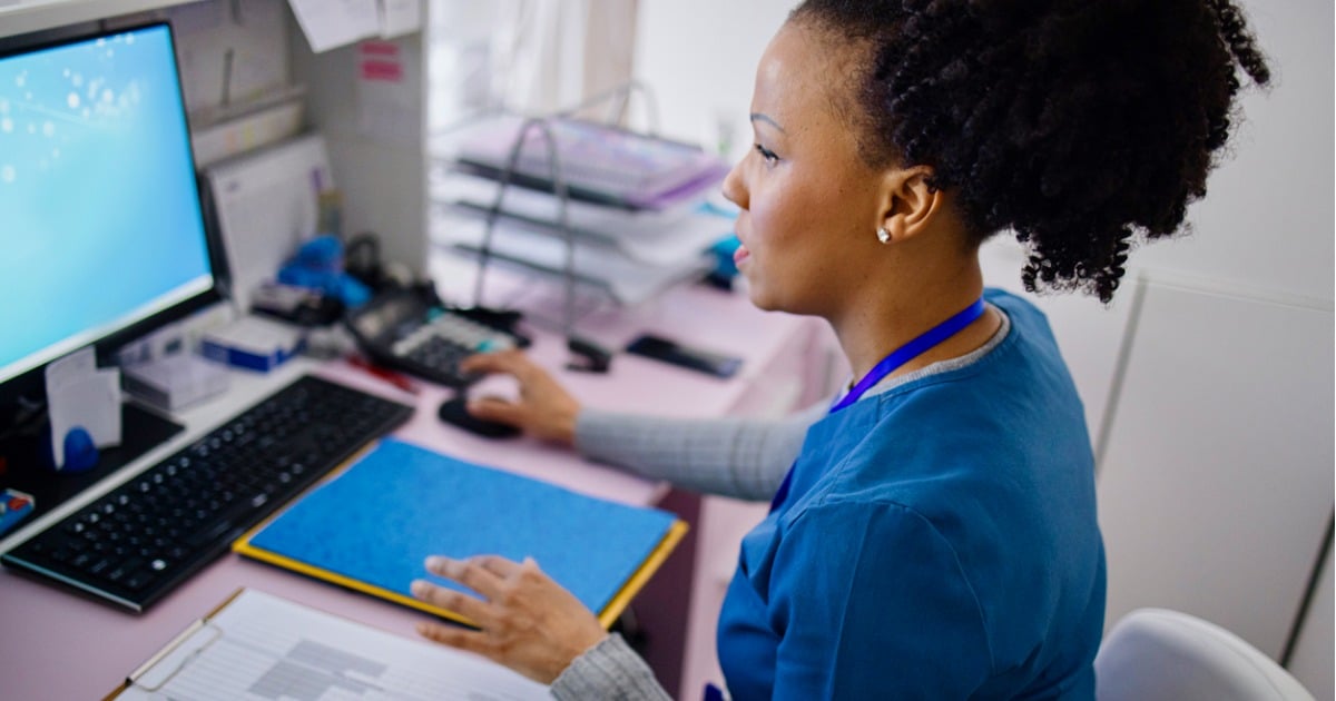 A woman in scrubs appears is looking at a computer screen and has her hand on the mouse. She appears to be at the front desk of a dental or medical clinic.