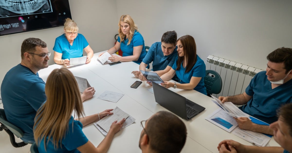 Dentists and dental team members in scrubs are seated around a table and appear to be reviewing documents during a meeting. A dental-related slide is projected on the wall behind them.